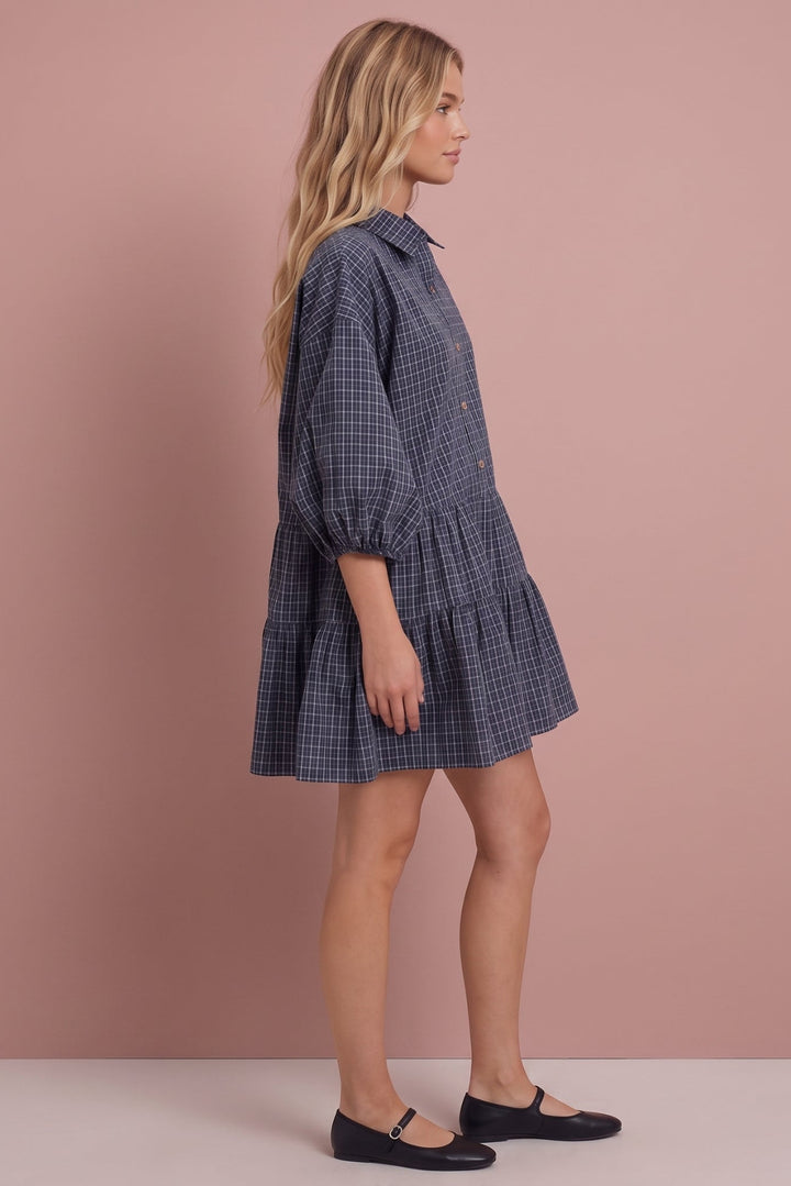 A woman modeling a navy and gray checkered mini shirt dress with puff sleeves, shown in profile against a solid dusty pink studio background.