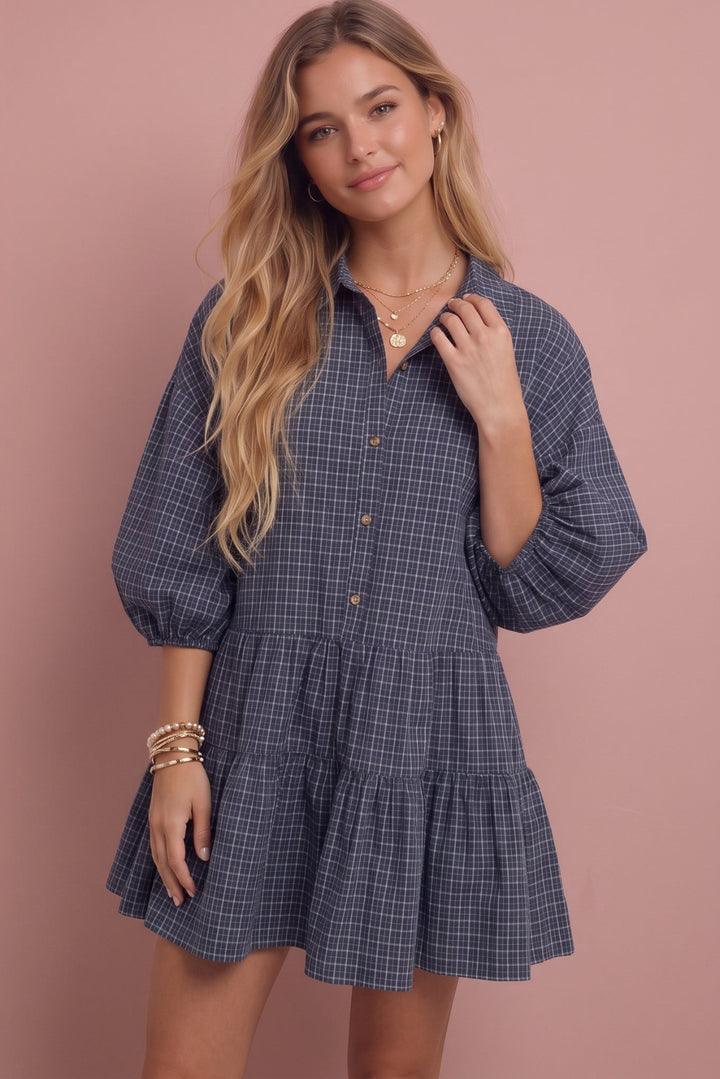 A woman modeling a blue and white plaid mini dress with puff sleeves and a tiered ruffle hem, posed against a solid dusty pink background.