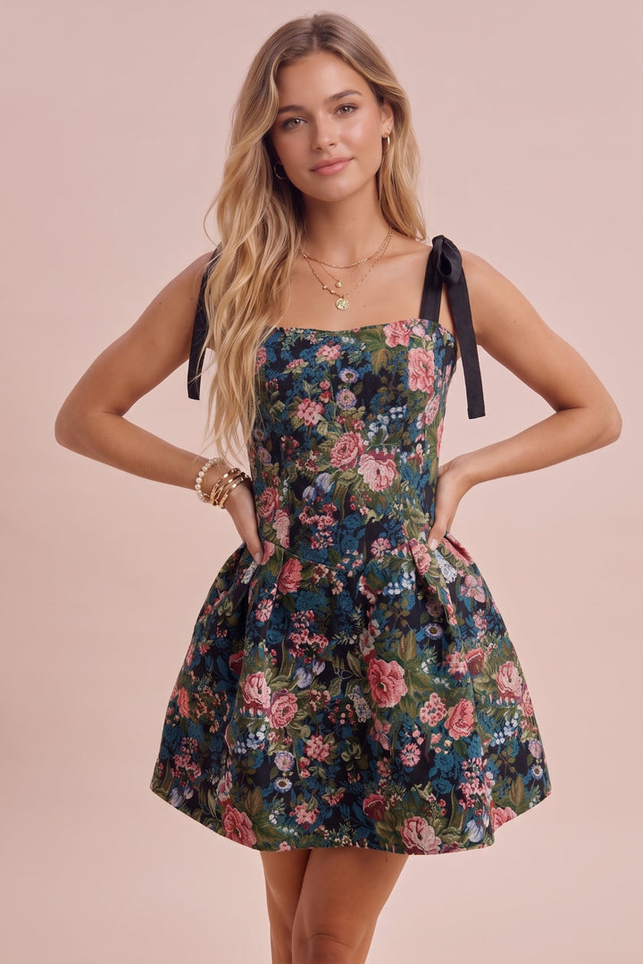 A woman modeling a sleeveless floral mini dress with black ribbon shoulder ties, standing against a solid pale pink background.