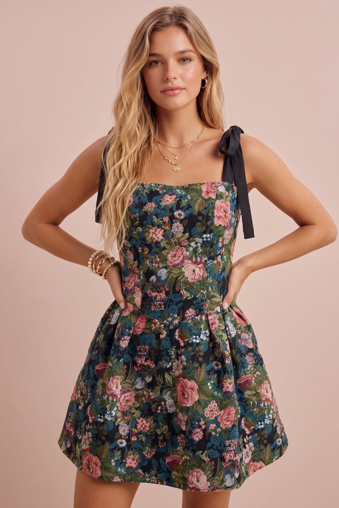 A woman modeling a sleeveless floral mini dress with black ribbon shoulder ties, standing against a solid pale pink background.