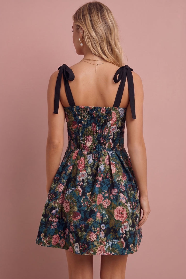 A woman shown from the back wearing a sleeveless floral mini dress with black ribbon tie straps against a solid dusty pink background.