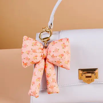 A fluffy, pink bow-shaped charm in a floral print pattern, attached to a white handbag or purse.