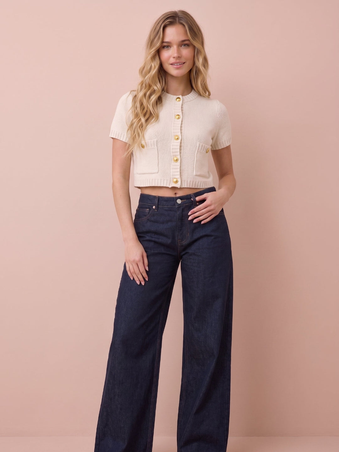A woman modeling a cream short-sleeve cropped cardigan with gold buttons and dark wide-leg jeans, standing against a solid pale pink background.