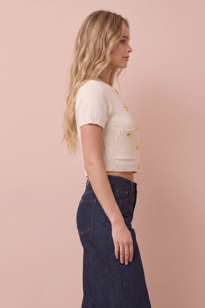 A woman in profile wearing a cream short-sleeve cropped knit top with gold buttons and dark wide-leg denim pants, standing against a solid dusty pink background.