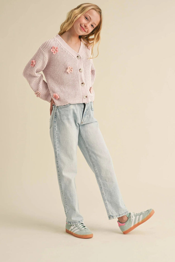 Young girl wearing a pink cardigan with floral embellishments, light blue jeans, and gray shoes on a beige background.