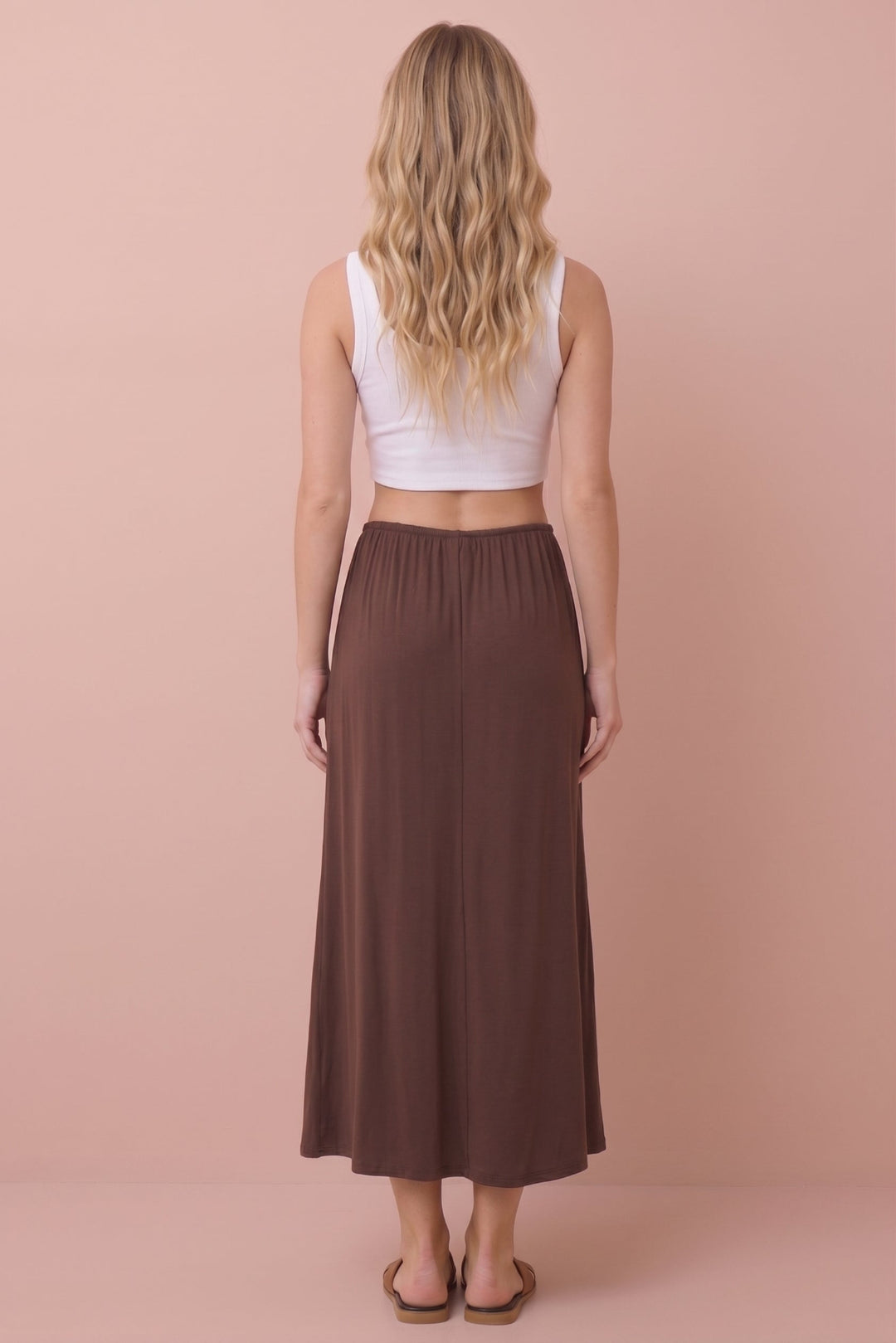 A woman shown from the back wearing a white sleeveless crop top and a long brown skirt, standing against a solid pale pink background.
