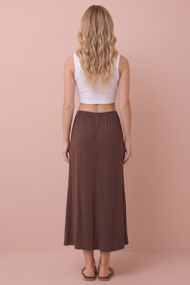 A woman shown from the back wearing a white sleeveless crop top and a long brown skirt, standing against a solid pale pink background.