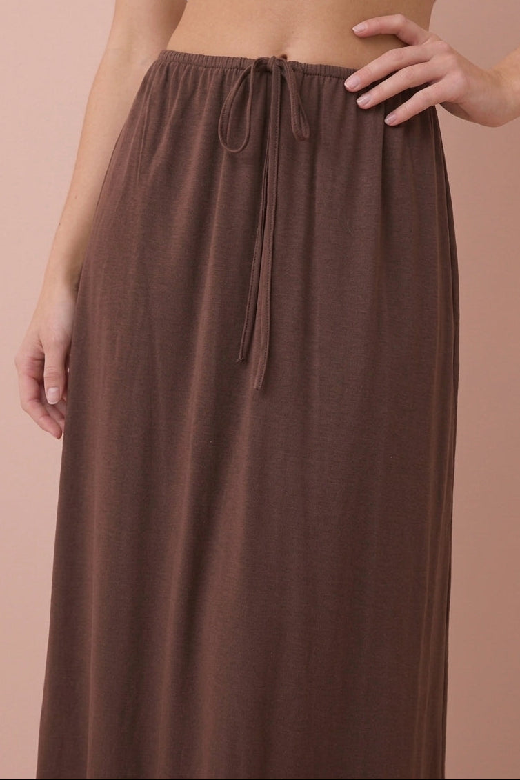 A model wearing a brown drawstring maxi skirt and a white crop top, standing against a solid peach-colored background.