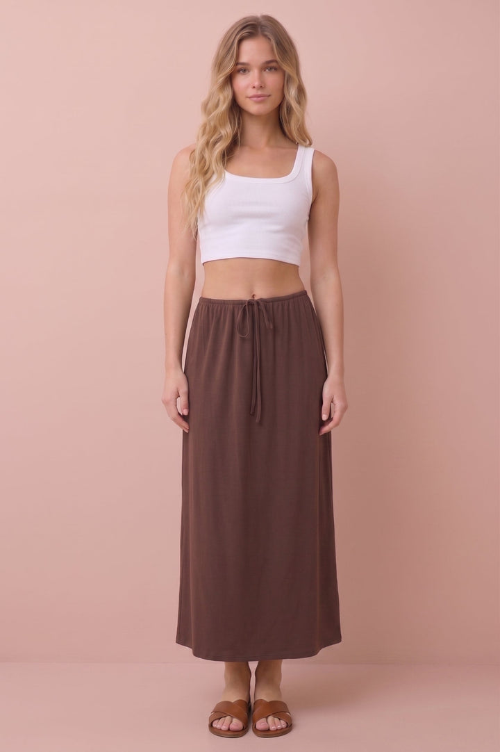 A woman stands against a solid dusty pink studio background wearing a white ribbed crop tank top and a long brown drawstring maxi skirt, paired with brown flat sandals.
