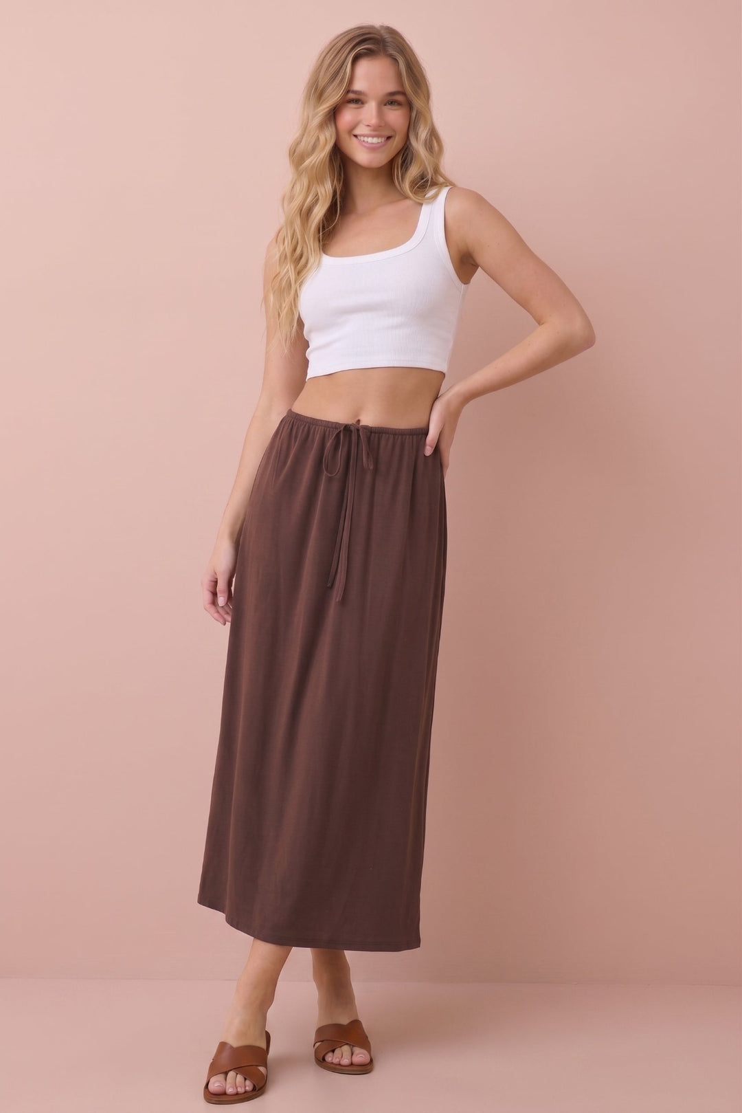 A woman modeling a brown midi skirt with a drawstring waist, paired with a white crop top and brown slide sandals, set against a solid dusty pink background.