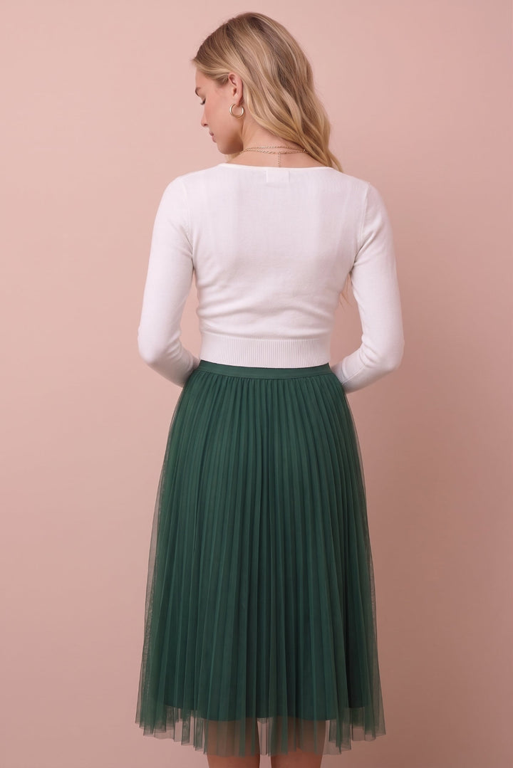 A woman shown from the back wearing a white long-sleeve knit top and a dark green pleated tulle midi skirt, standing against a solid dusty pink background.