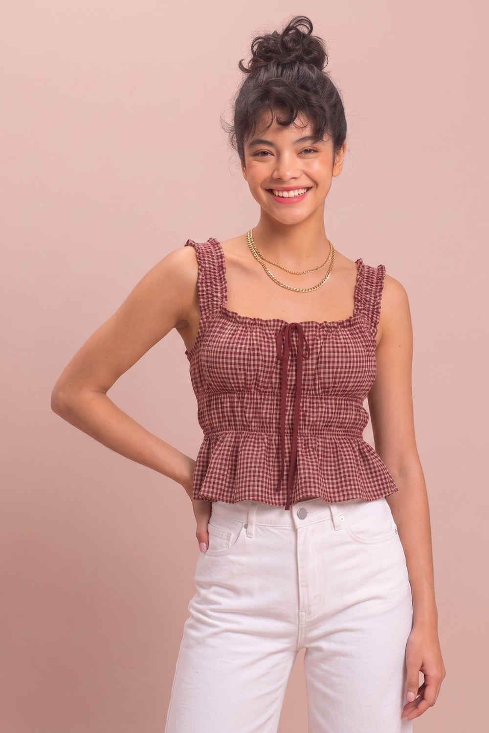 A woman modeling a sleeveless red and white gingham peplum top with a ruffled neckline and front tie, paired with white pants, against a solid dusty pink background.