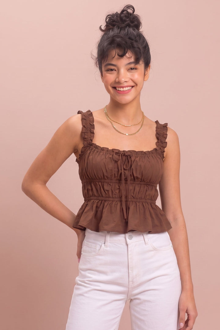 A woman modeling a brown ruffled spaghetti-strap top with a tie detail at the neckline, paired with white pants, against a solid light pink background.