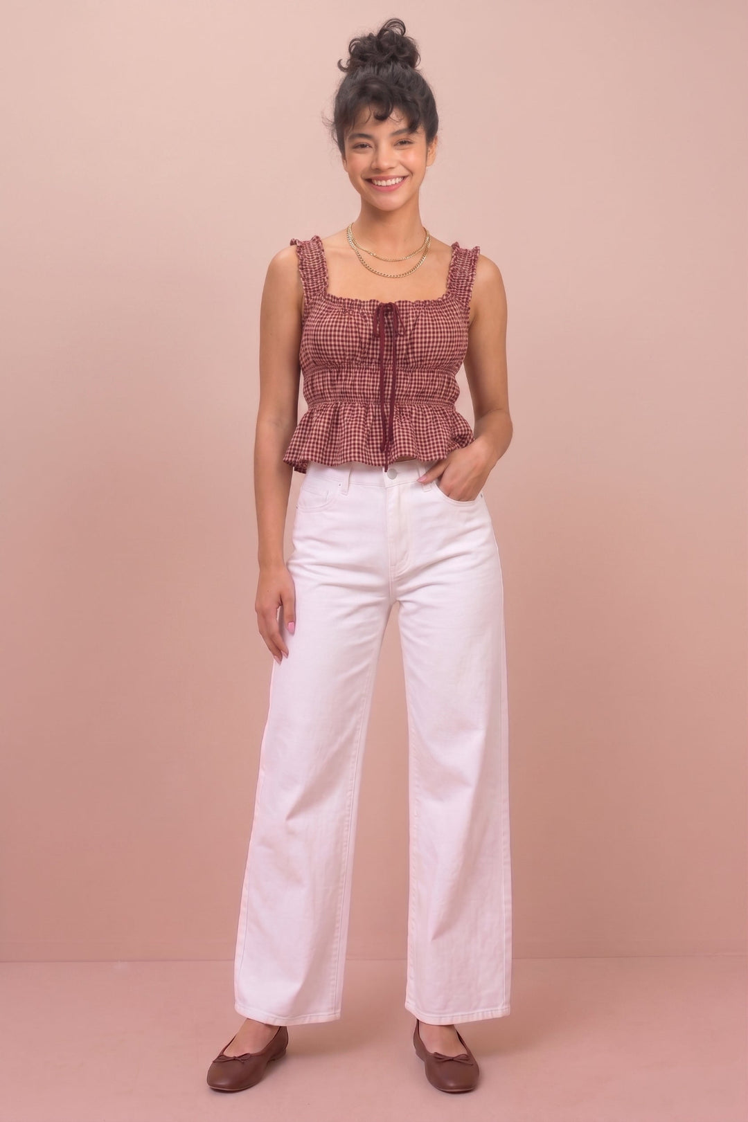 A woman modeling a sleeveless ruffled top in a muted reddish-brown and white gingham pattern, paired with white wide-leg pants and brown flats, standing against a solid dusty pink background.