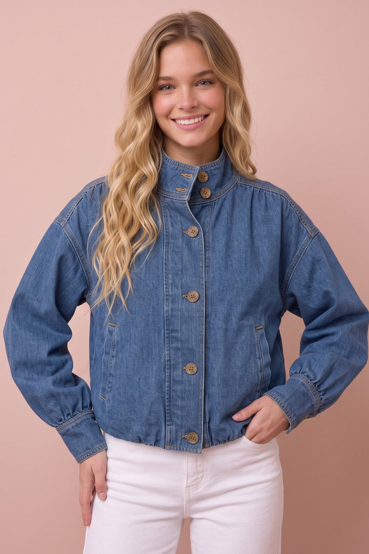 A woman modeling a cropped blue denim jacket with a high stand collar and brown buttons, paired with white pants, set against a solid dusty pink background.