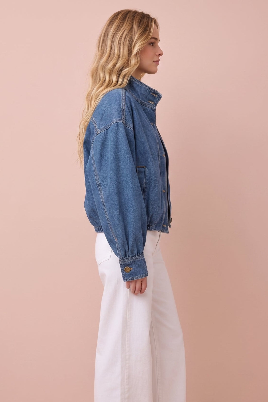 A woman stands in profile wearing a cropped blue denim jacket, white wide-leg pants, and brown slip-on shoes against a solid pale pink studio background.