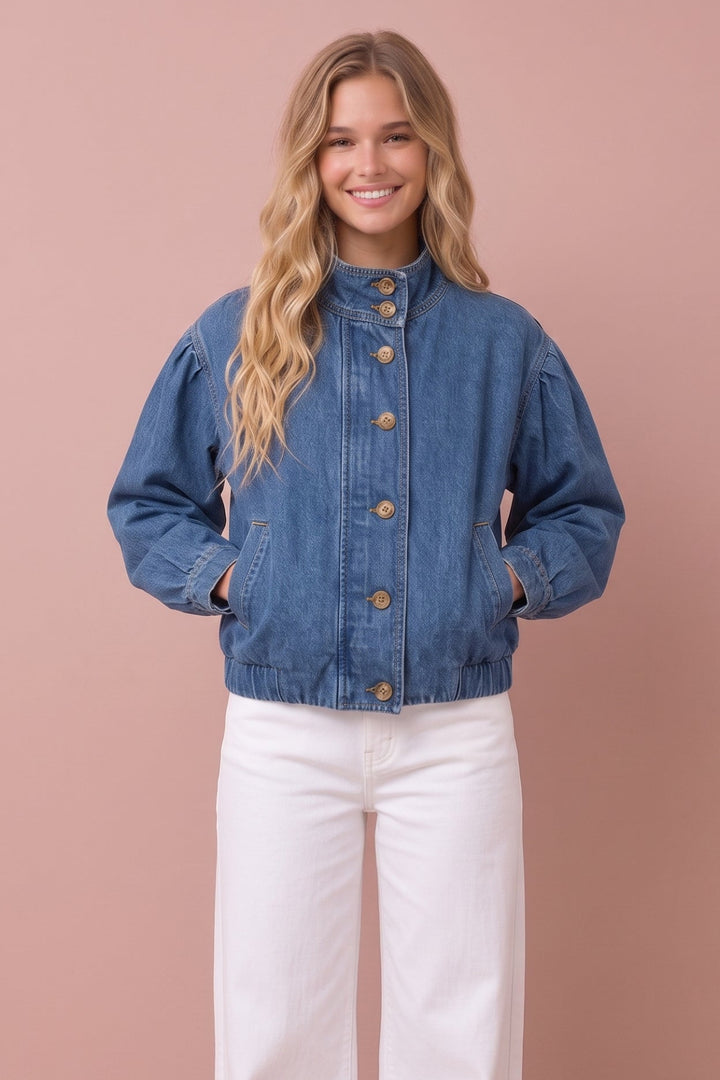 A woman modeling a blue denim jacket with a high mock neck and brown buttons, paired with white wide-leg pants, against a solid dusty pink background.