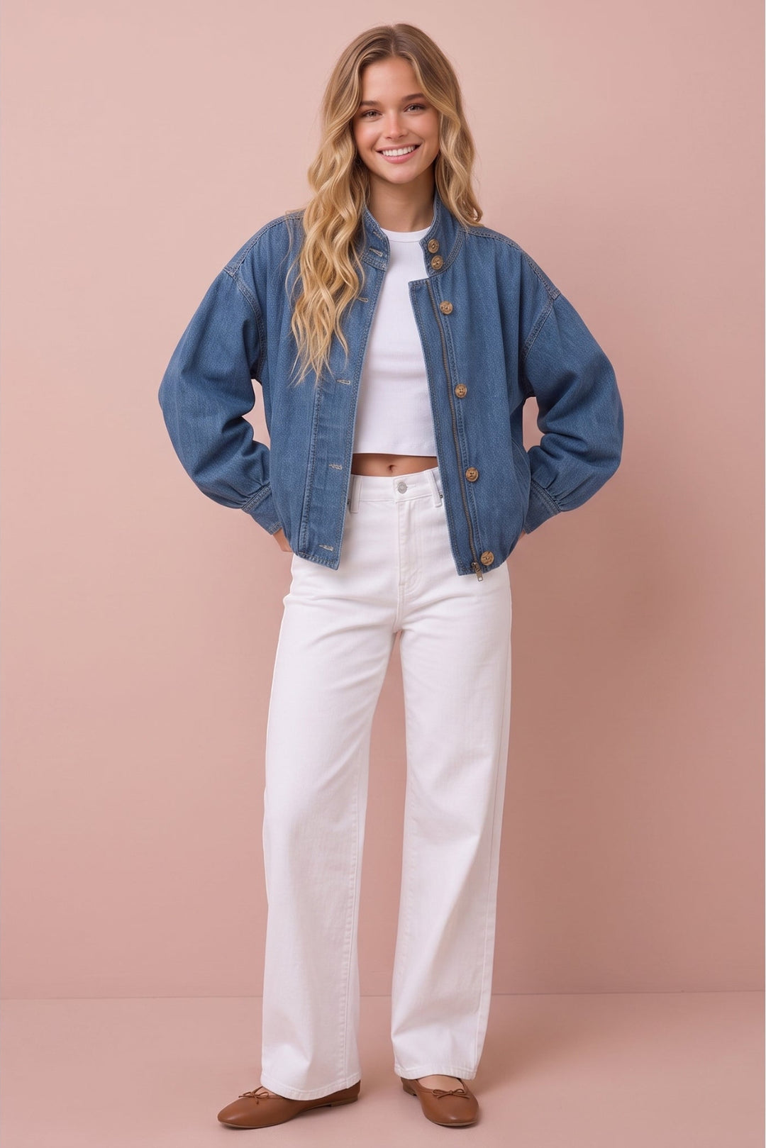 A woman stands against a dusty pink studio backdrop wearing a blue denim jacket with brown buttons over a white crop top, white wide-leg pants, and brown flats.
