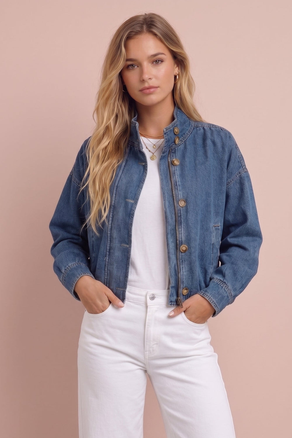 A woman modeling a blue denim jacket over a white top and white wide-leg jeans, standing against a solid pale pink background.