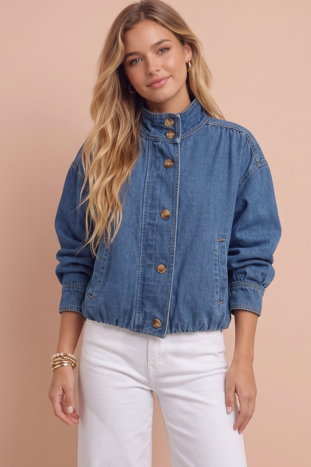 A woman modeling a cropped blue denim jacket with a high stand collar and brown buttons, paired with white pants, against a solid peach background.