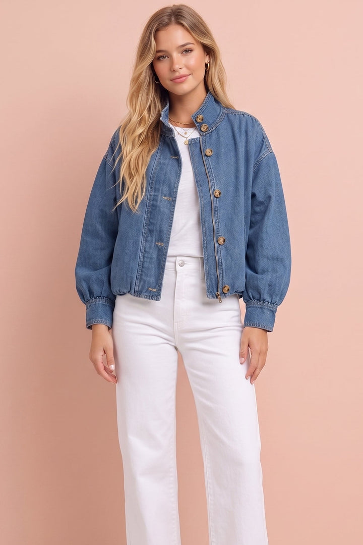 A woman modeling a cropped blue denim jacket with buttoned cuffs and hem, worn over a white top and white wide-leg jeans, standing against a solid peach-pink studio background.