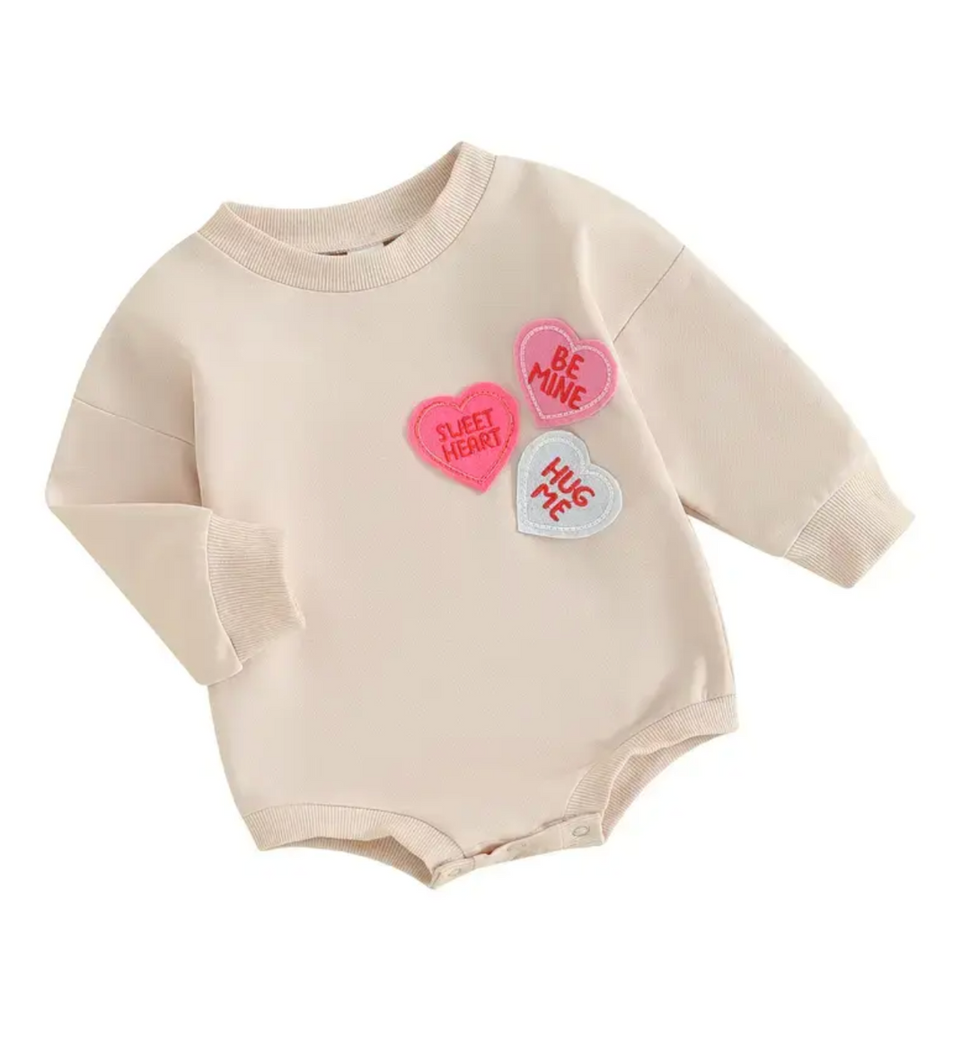 A cream-colored baby sweatshirt romper with pink and gray heart-shaped patches reading "Sweet Heart" and "Be Mine".