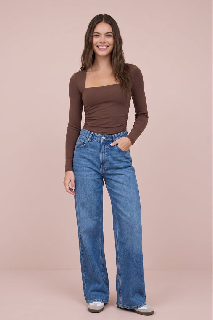A woman modeling a brown long-sleeve square-neck top and high-waisted, wide-leg blue jeans against a solid pale pink studio background.