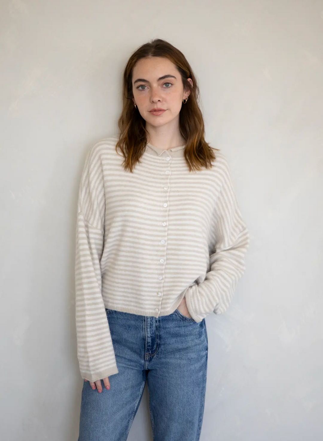 A young woman wearing a beige and white striped knit cardigan, standing against a white wall. The cardigan has a button-up front and long sleeves.