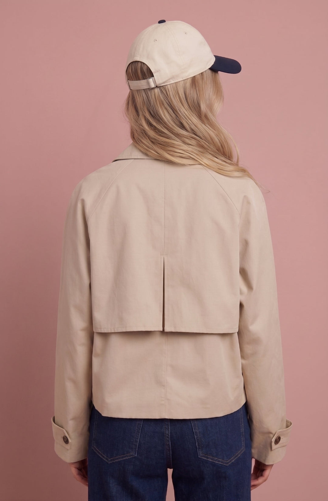 Cream cropped trench jacket worn with dark jeans, back view on pink background.