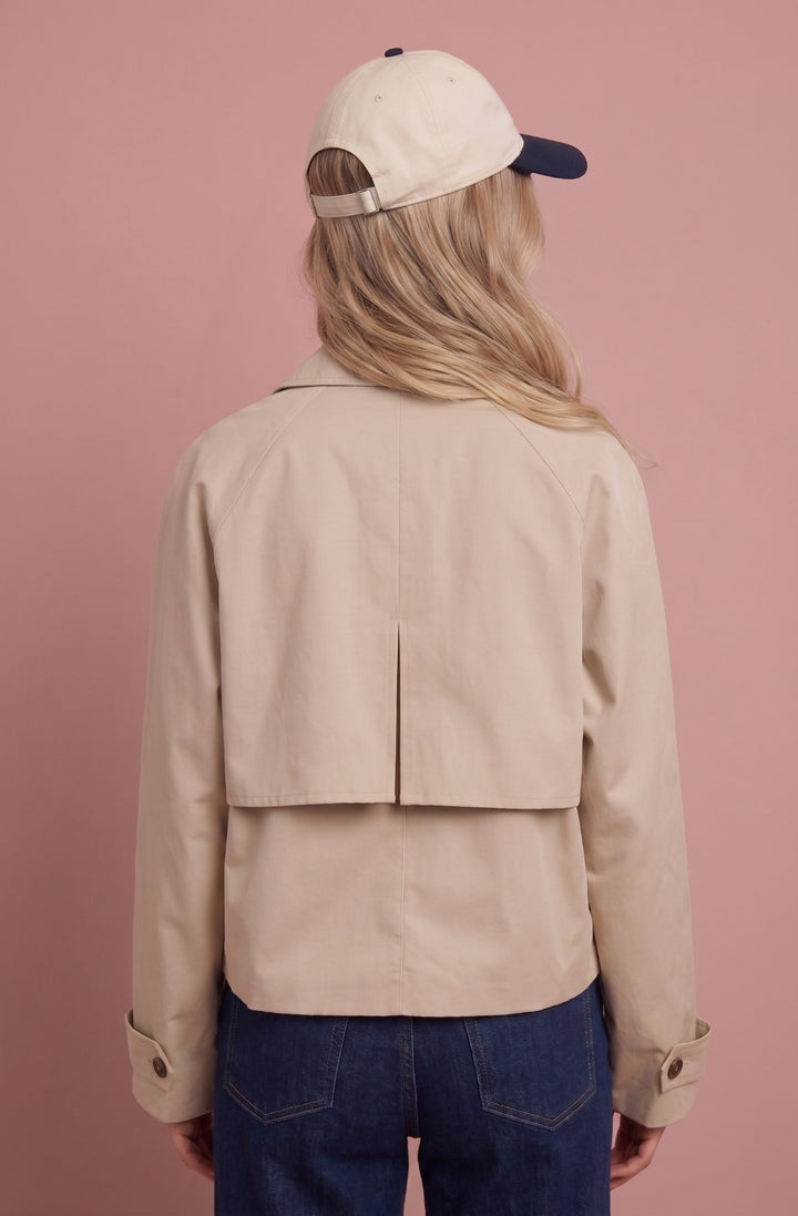 Cream cropped trench jacket worn with dark jeans, back view on pink background.