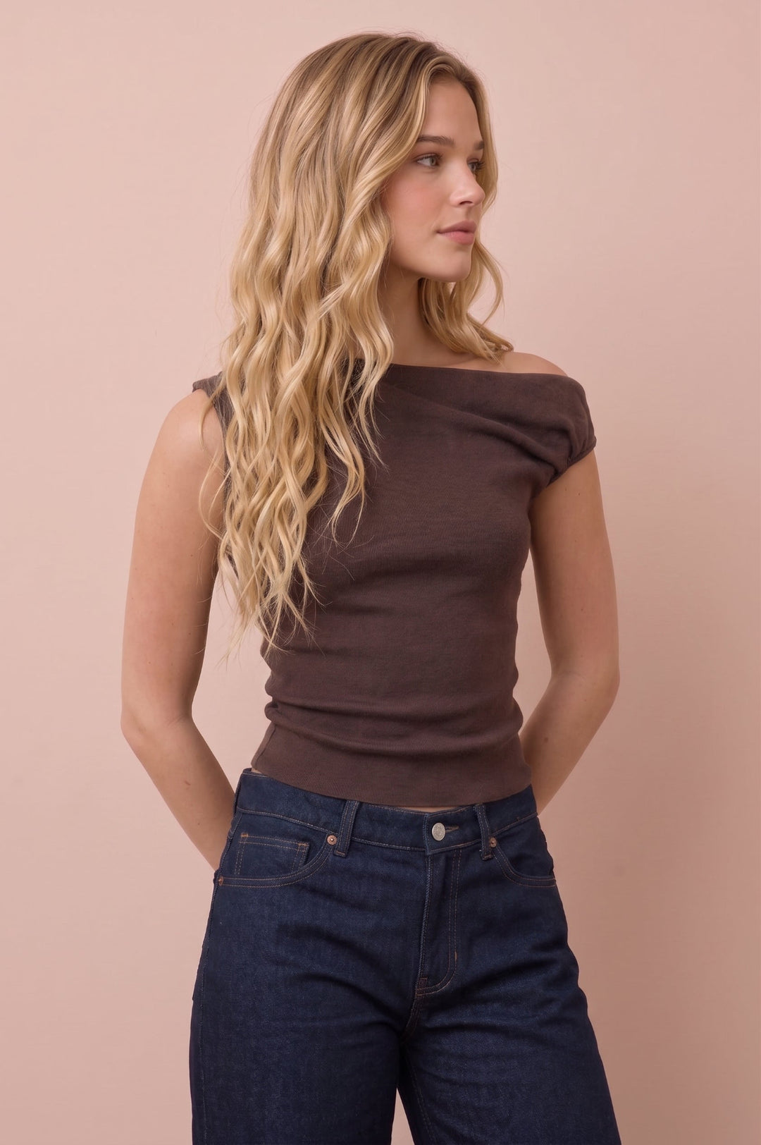 A woman with long wavy blonde hair wearing a dark brown off-the-shoulder ribbed top and dark blue jeans, standing against a solid pale pink background.