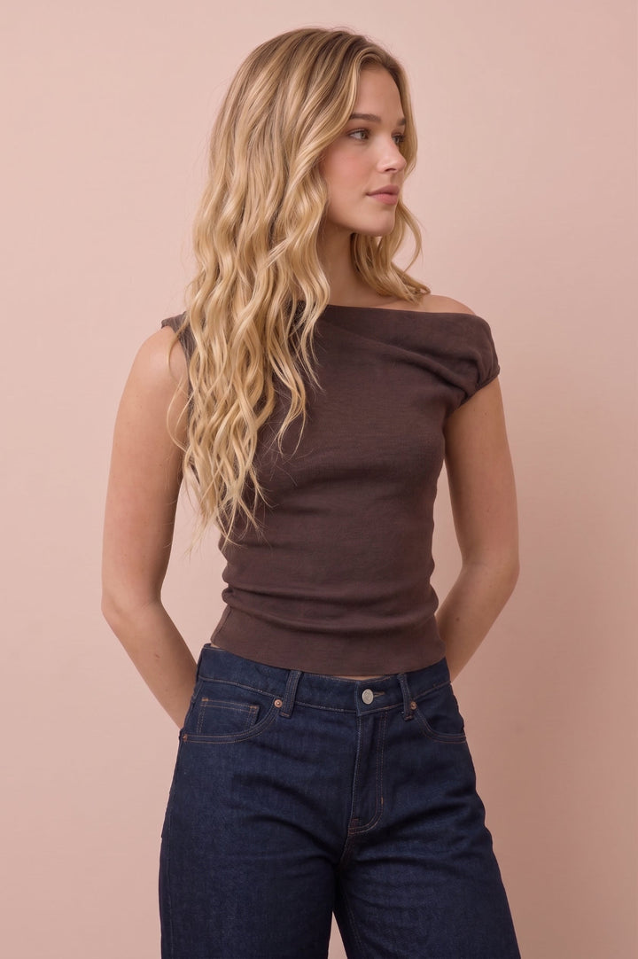 A woman with long wavy blonde hair wearing a dark brown off-the-shoulder ribbed top and dark blue jeans, standing against a solid pale pink background.