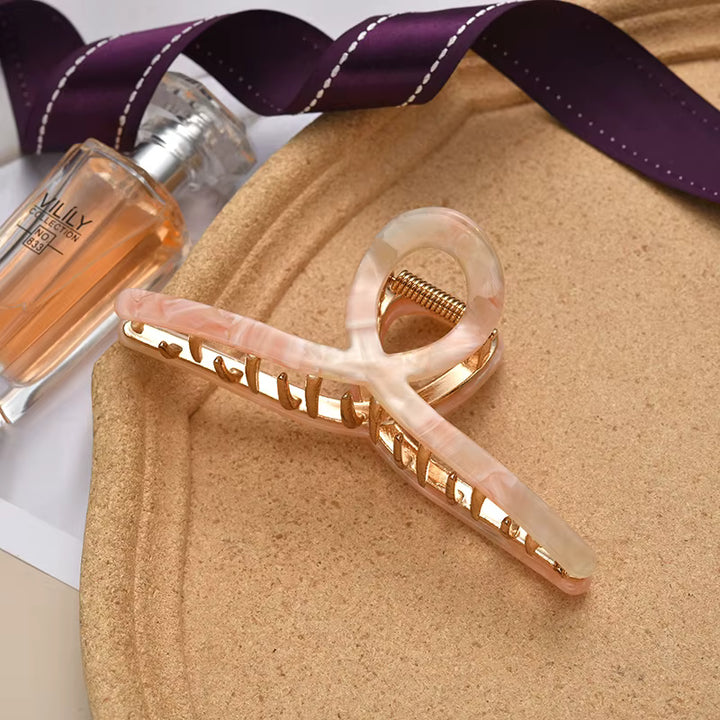 A gold-toned claw hair clip with a scalloped design, set against a purple satin ribbon and perfume bottle.