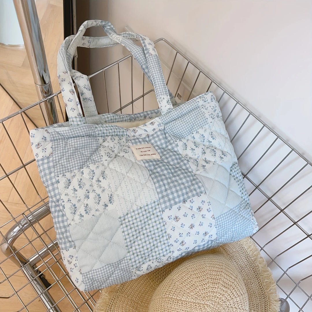 A patchwork quilted tote bag in muted blue and white gingham and floral patterns, with a zipped pocket and straps for carrying.