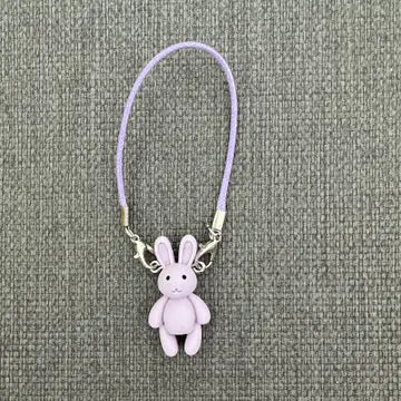 A lavender-colored plush bunny figurine attached to a thin purple cord or strap, used as an accessory for handmade dolls.