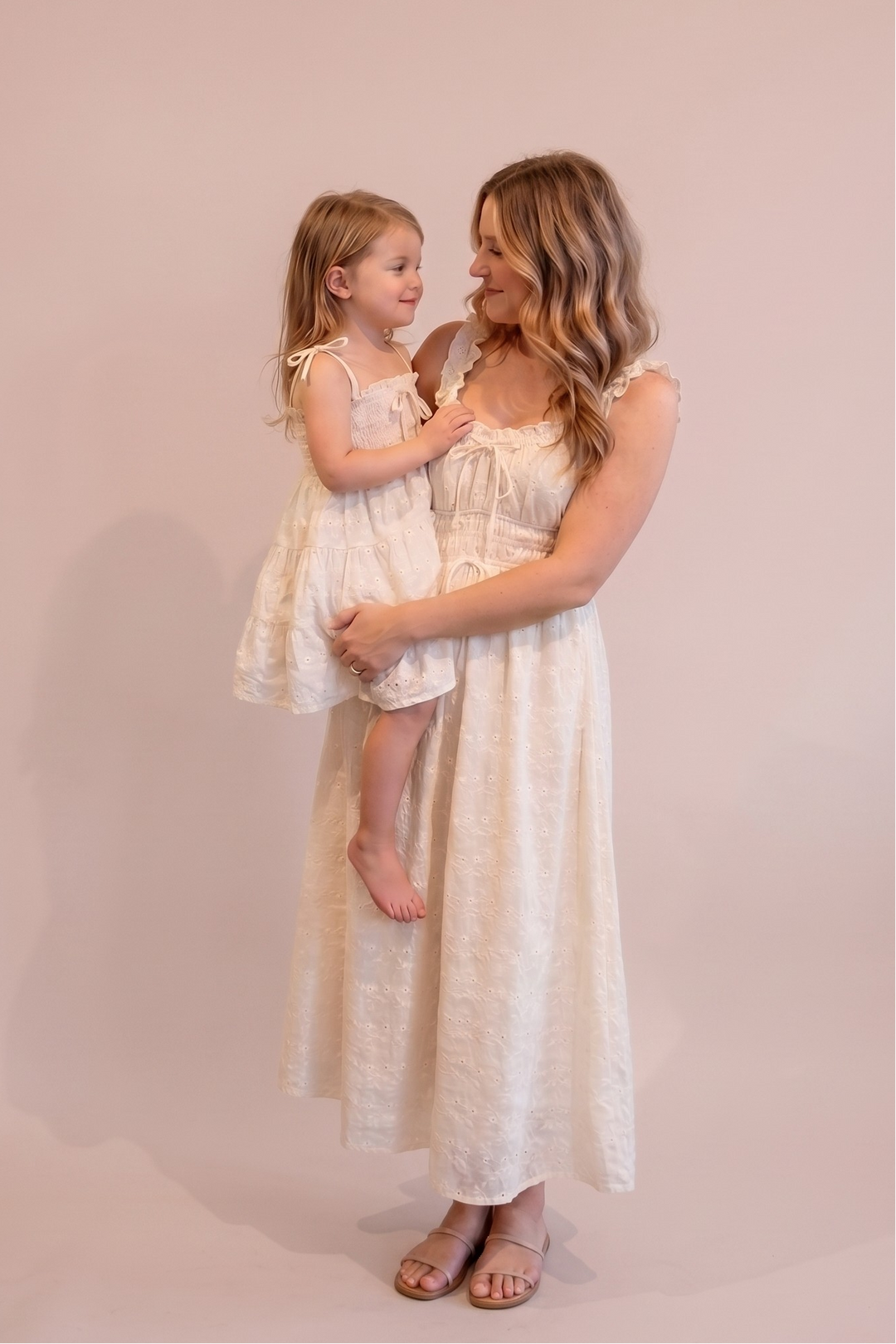 A woman and a young girl wearing matching cream-colored textured dresses pose together against a solid light pink studio backdrop.