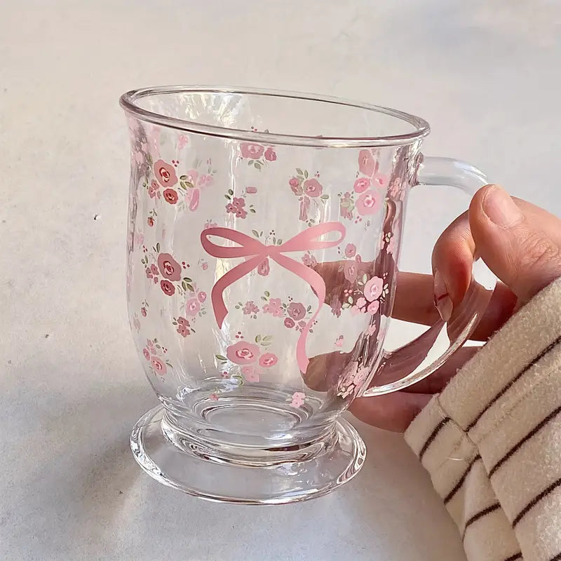 A transparent glass mug with delicate pink floral patterns and a pink ribbon bow decoration.