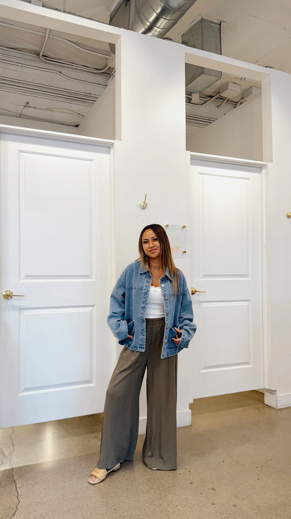 Woman standing in a room with white walls and doors, wearing a denim jacket and gray pants.