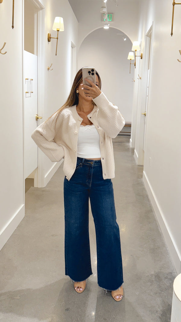Woman taking a mirror selfie wearing a white cardigan, white top, and blue jeans in a hallway.