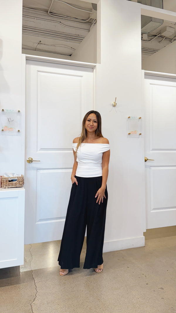 Woman in a white off-shoulder top and black wide-leg pants standing in a room with white walls and doors.