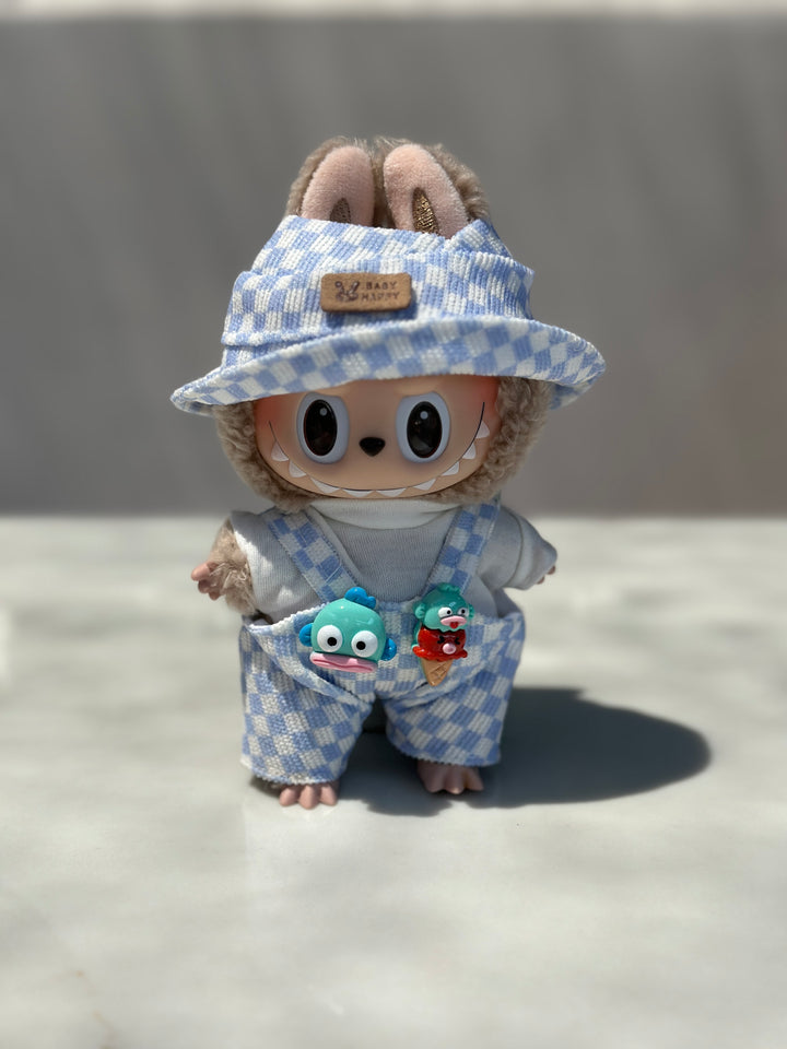 A plush toy doll wearing blue and white checked overalls with a matching bucket hat, adorned with colorful monster charms.