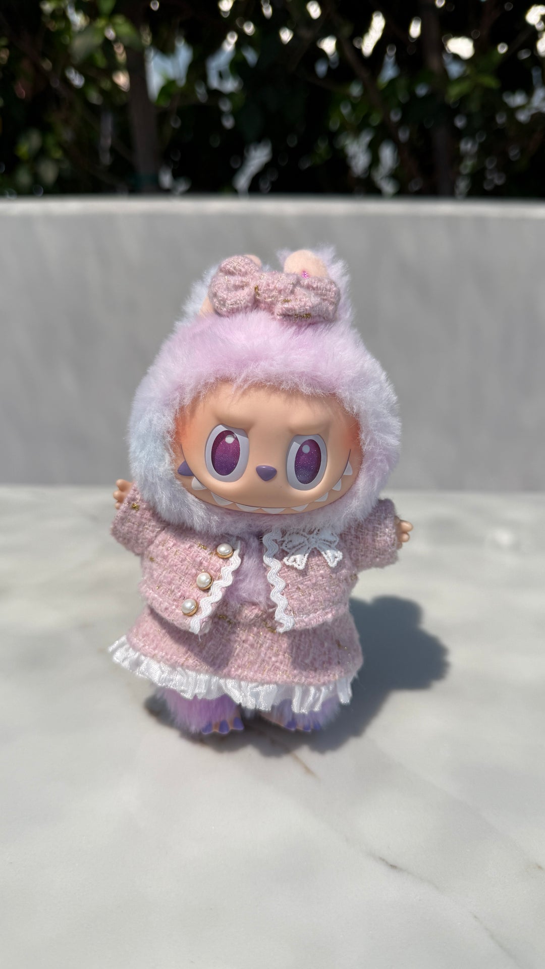 A fluffy, lavender-colored plush doll wearing a ruffled pink jacket with a bow headpiece.