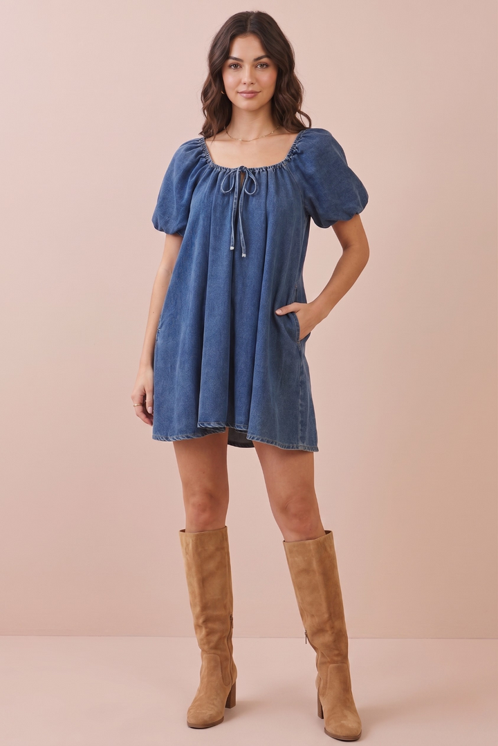 A woman modeling a short-sleeve blue denim dress with puff sleeves, a square neckline, and a drawstring tie, paired with tan knee-high boots against a solid pale pink studio background.