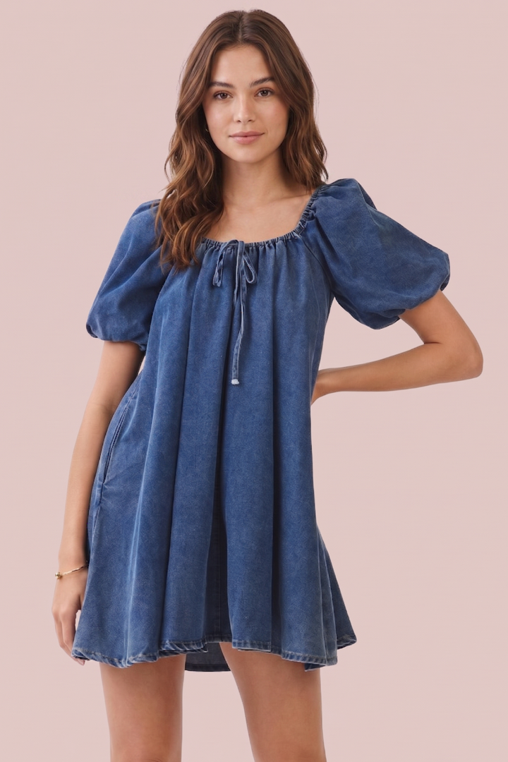 A woman modeling a blue denim puff-sleeve mini dress with a tie neckline, set against a solid pale pink background.
