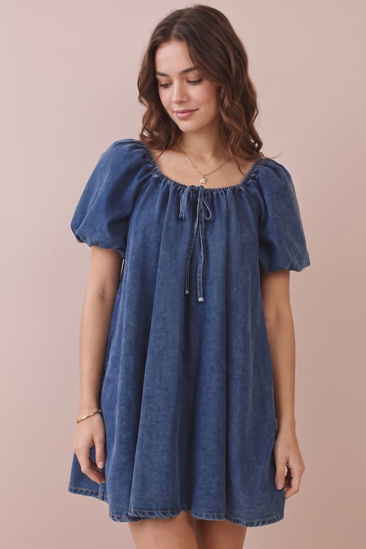 A woman wearing a blue denim dress with puff sleeves, a square neckline, and a front tie detail, posed against a solid light pink background.