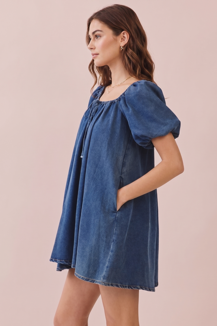 A woman wearing a blue denim puff-sleeve mini dress, shown from a side profile against a solid pale pink background.