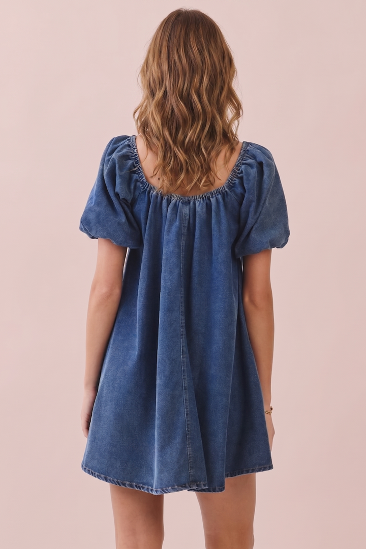 A woman viewed from the back wearing a blue denim dress with short puff sleeves and a square neckline, standing against a solid pale pink background.