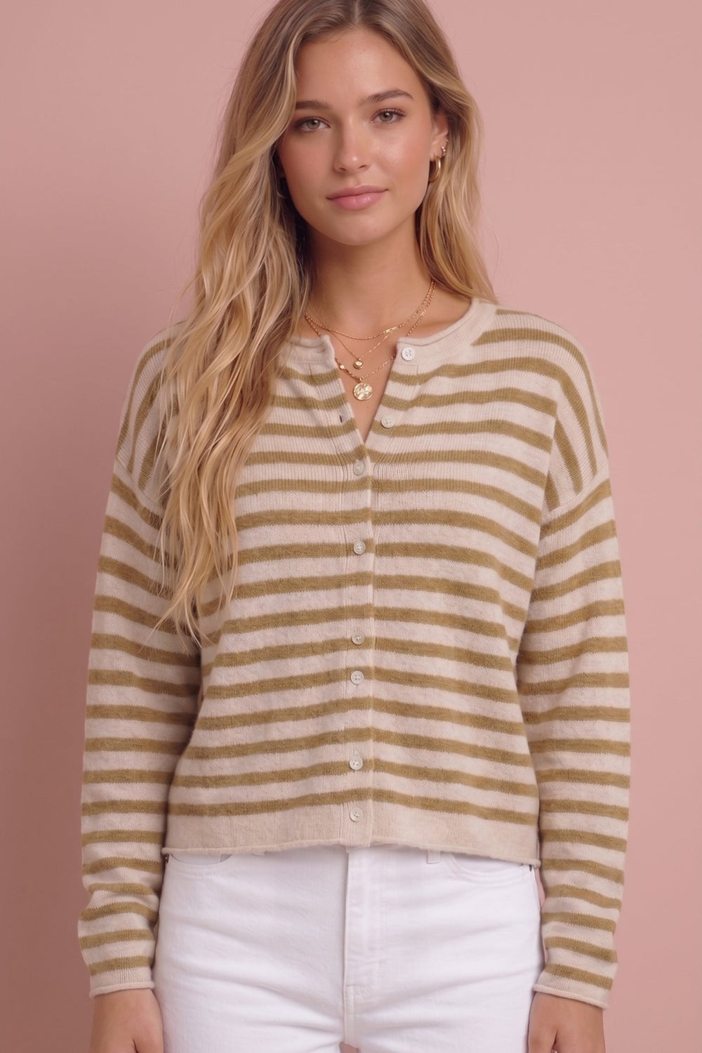 A woman modeling a cropped, long-sleeve knit cardigan with horizontal tan and cream stripes, featuring a split neckline with button details, paired with white pants against a solid dusty pink background.