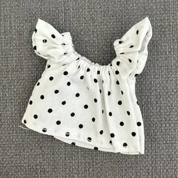 White sleeveless shirt with black polka dots, featuring ruffled shoulders and an oversized bow detail.