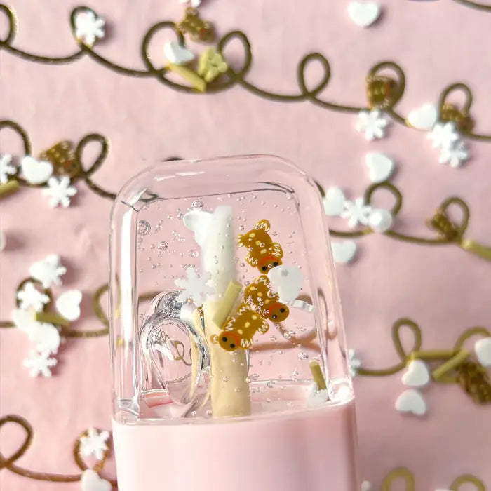 A glass dome containing a white popsicle-shaped lip gloss with gold glitter accents, set against a pink background with gold floral illustrations.